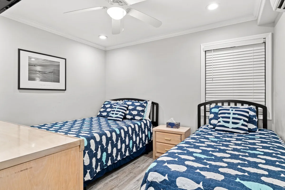 A comfortable bedroom with two beds dressed in nautical-themed bedding, framed artwork on the wall, and a small nightstand with a lamp. Morning light filters through, creating a welcoming and peaceful space for families or friends to relax together.
