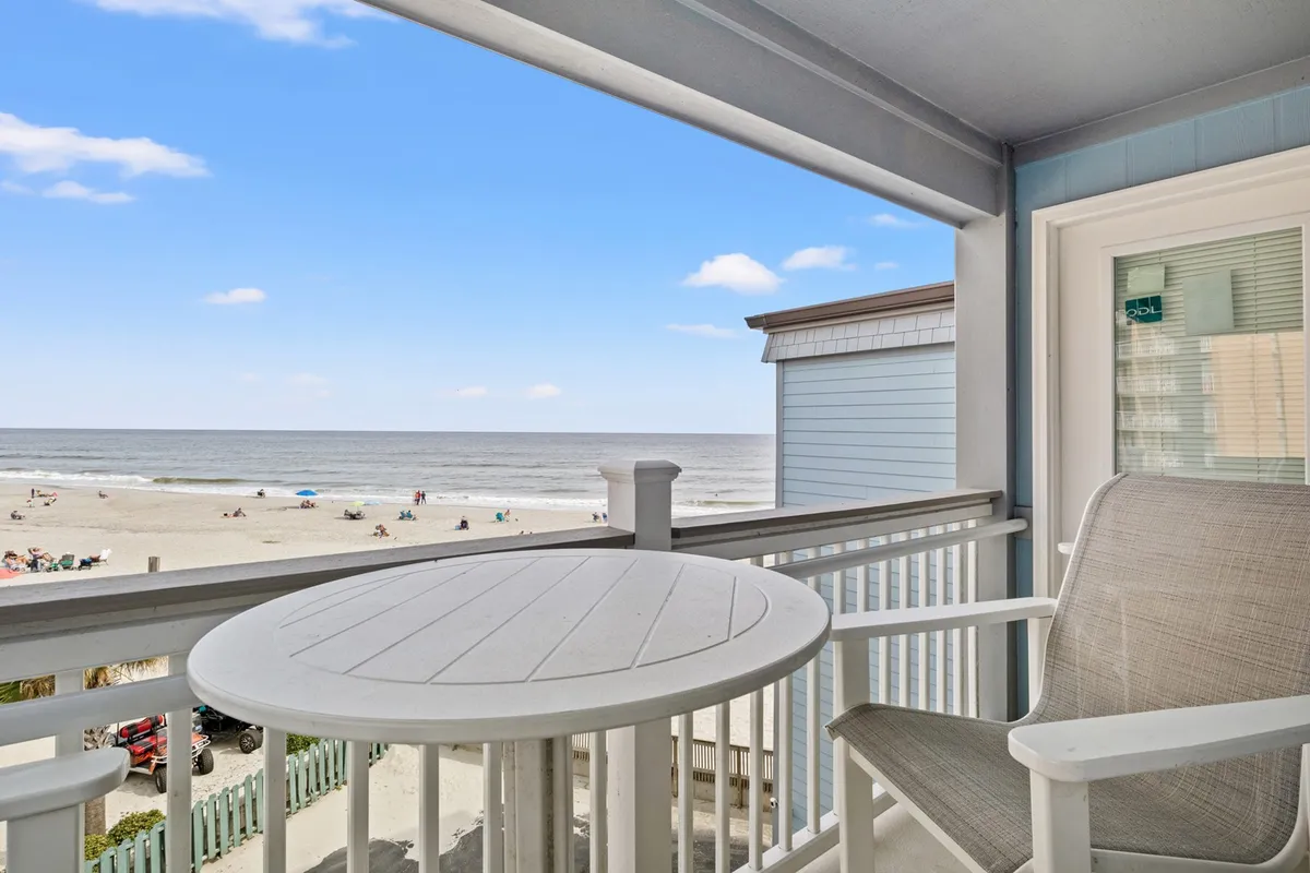 The image shows a private condo balcony with inviting seating, overlooking a pristine stretch of Myrtle Beach. Beyond the railing, gentle waves roll onto the sun-kissed shore dotted with beachgoers, all set against a backdrop of endless blue sky and sparkling ocean.