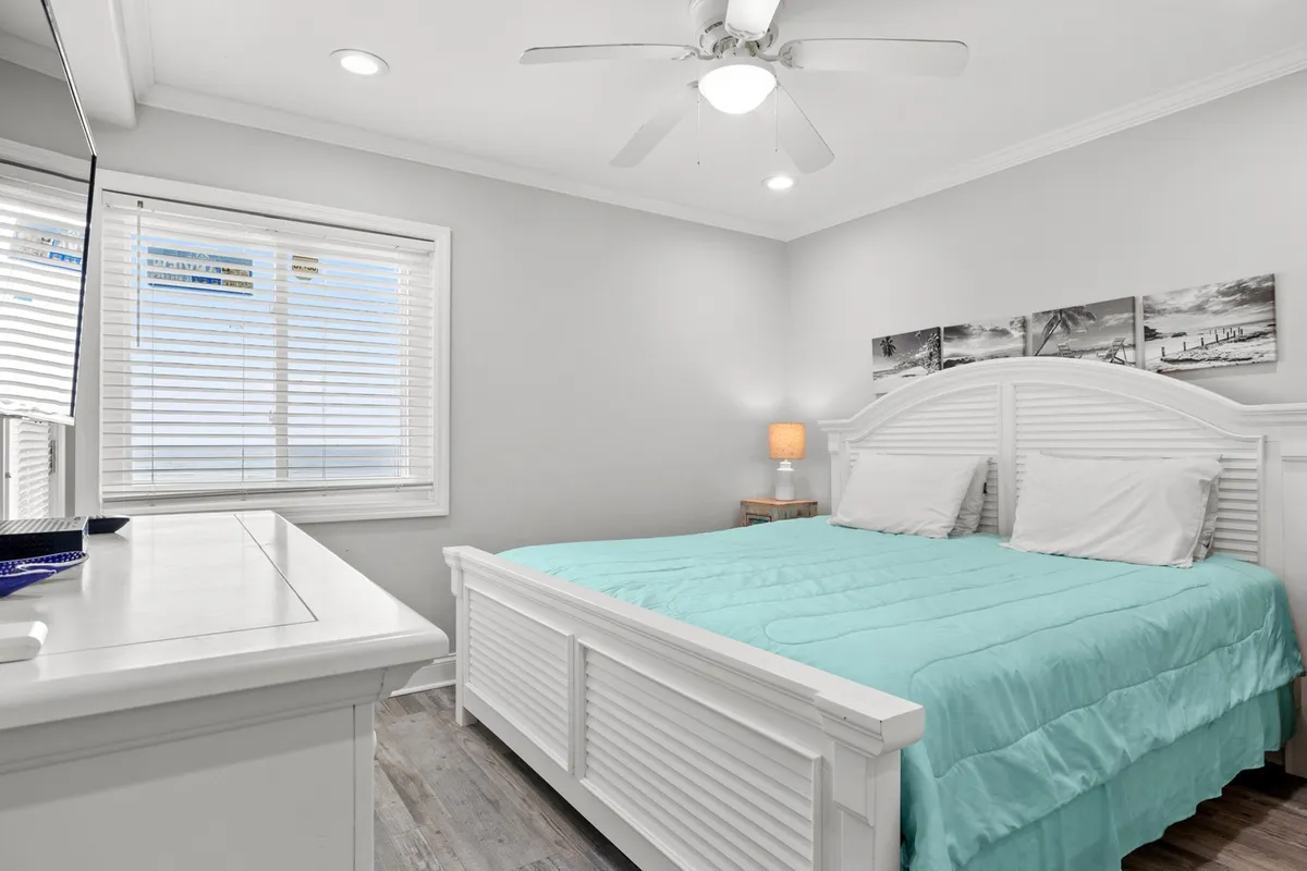 A tranquil bedroom features a bed with plush turquoise bedding, a wooden nightstand, and decorative photographs on the wall. A ceiling fan overhead and soft natural light contribute to the relaxing atmosphere, inviting restful moments.