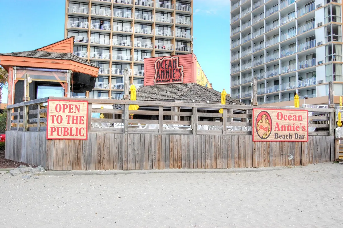 This image captures the iconic Ocean Annie's Beach Bar with its eye-catching sign, wooden fence, and a lively outdoor setting featuring yellow umbrellas. Guests are seen enjoying drinks, socializing, and relaxing right next to the beach. The scene is lit with the warm hues of sunset, highlighting the venue's fun and vibrant atmosphere that comes alive with live music and themed parties.