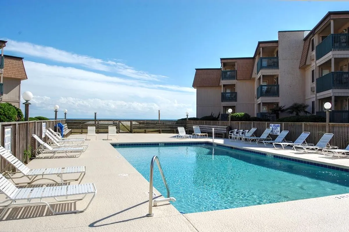 A spacious outdoor pool area bordered by inviting lounge chairs and a backdrop of condominium buildings. The pool deck is perfect for sunbathing or relaxing after a swim, with plenty of space for social gatherings and family fun.
