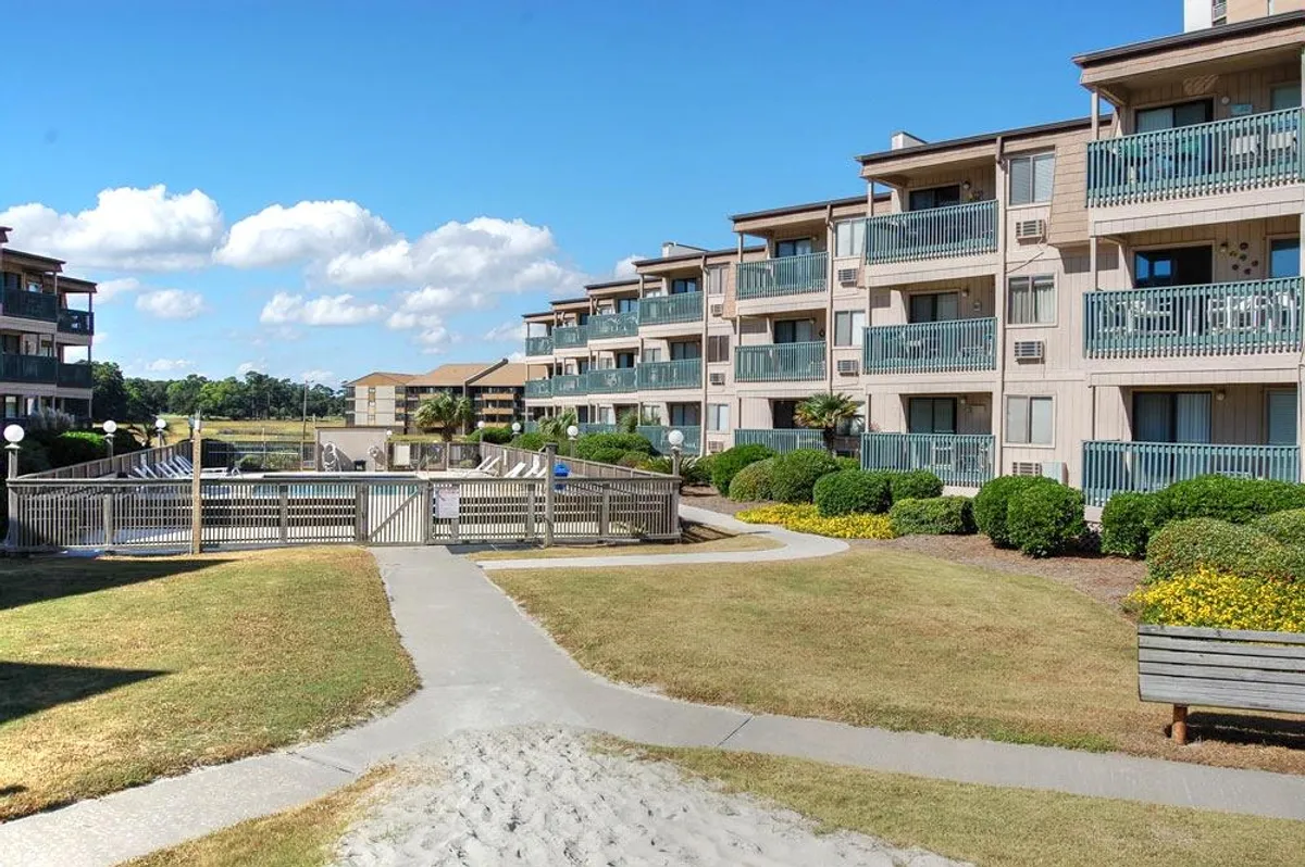 A landscaped exterior with balconies, gardens, a well-maintained walkway, and a gated pool area in view. The inviting pathways connect various resort features, including sun decks and communal spaces for relaxation or gatherings.