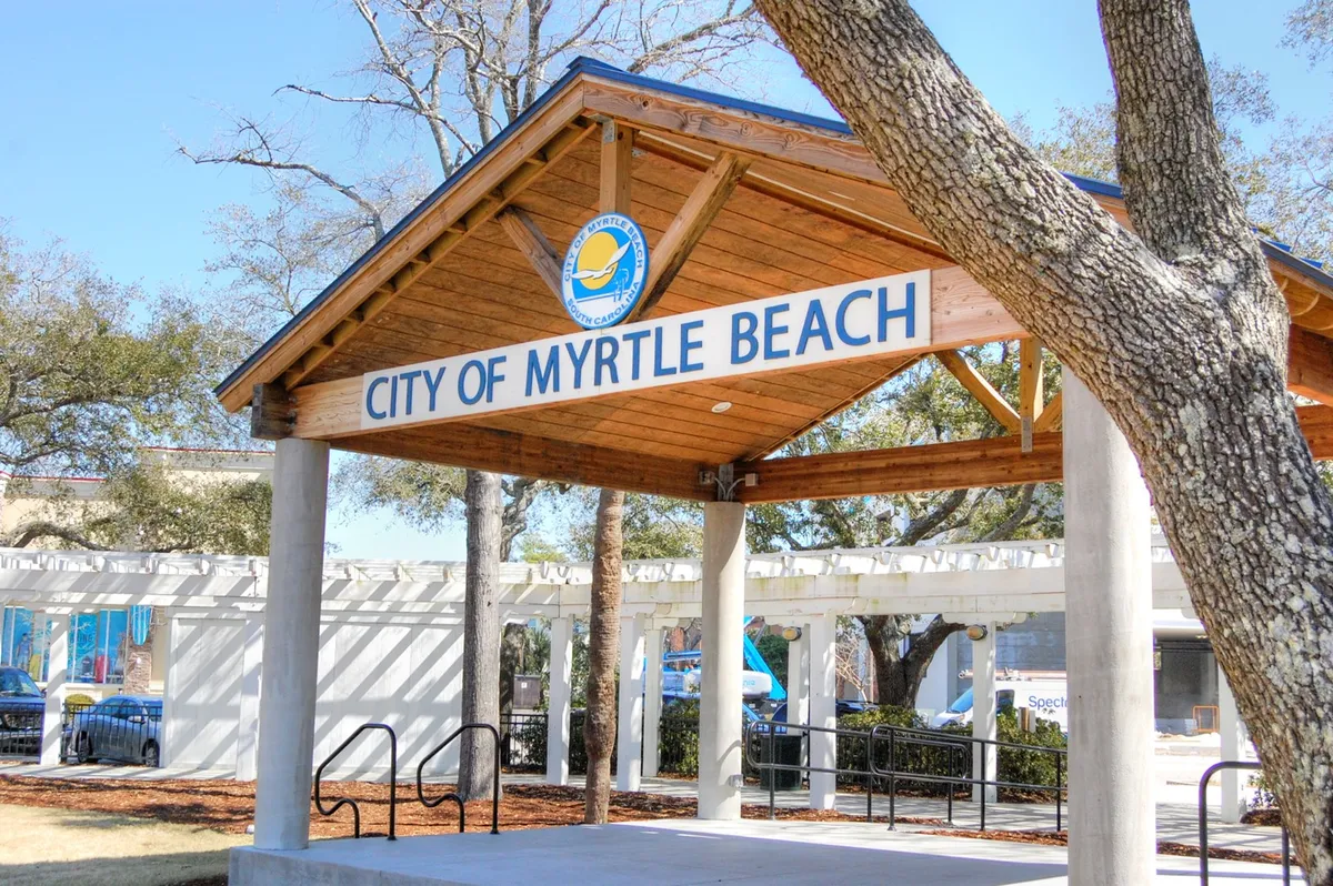A welcoming wooden pavilion bearing the 'CITY OF MYRTLE BEACH' sign, framed by lush green trees. The area is filled with natural light and set up for community gatherings or relaxed afternoons.