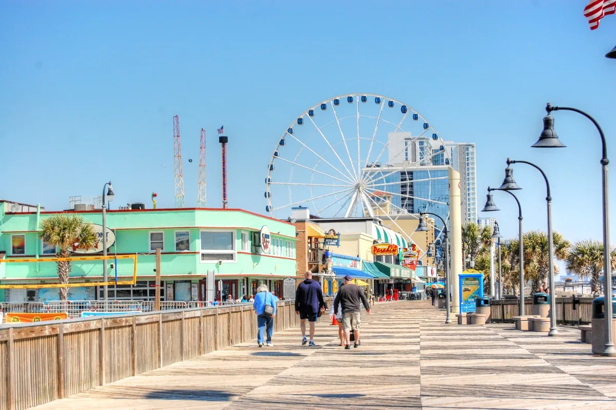 A lively boardwalk scene featuring a ferris wheel, colorful storefronts, and people strolling along the wooden planks. The atmosphere is festive and energetic, evoking the spirit of seaside vacations and community events.